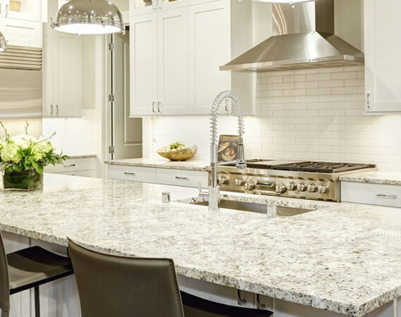 White kitchen design features large bar style kitchen island with granite countertop illuminated by modern pendant lights. Stainless steel appliances framed by white shaker cabinets . Northwest, USA